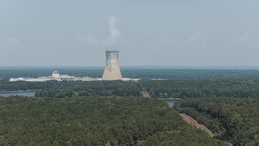 A wide, slow-panning aerial shot of a nuclear power plant and its cooling tower releasing steam, surrounded by a vast forest and lake