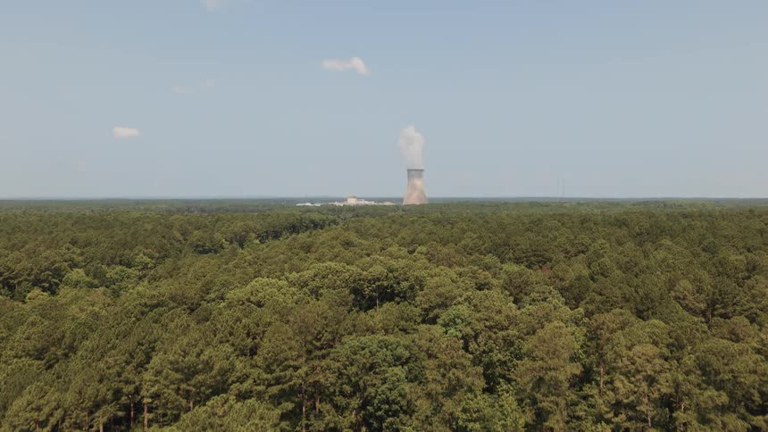 An aerial drone shot rises above a dense green forest to reveal a nuclear power plant and cooling tower on the horizon