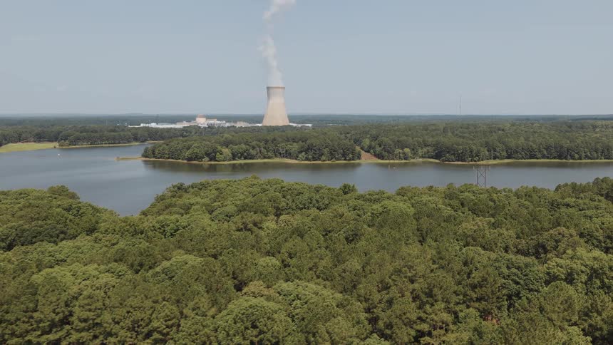 Slow aerial retreat over a lush green forest revealing a nuclear power plant and its cooling tower steaming beside a large lake