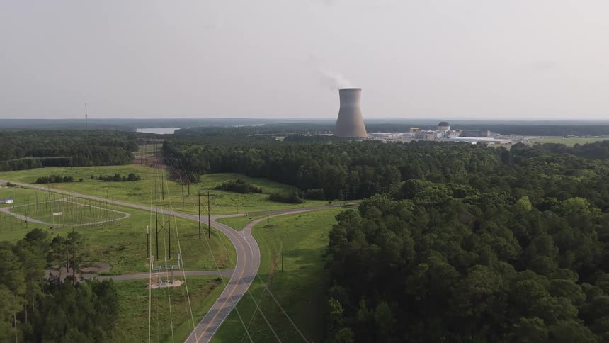 Slow aerial shot moving towards a nuclear power plant and its cooling tower surrounded by a dense green forest and power lines