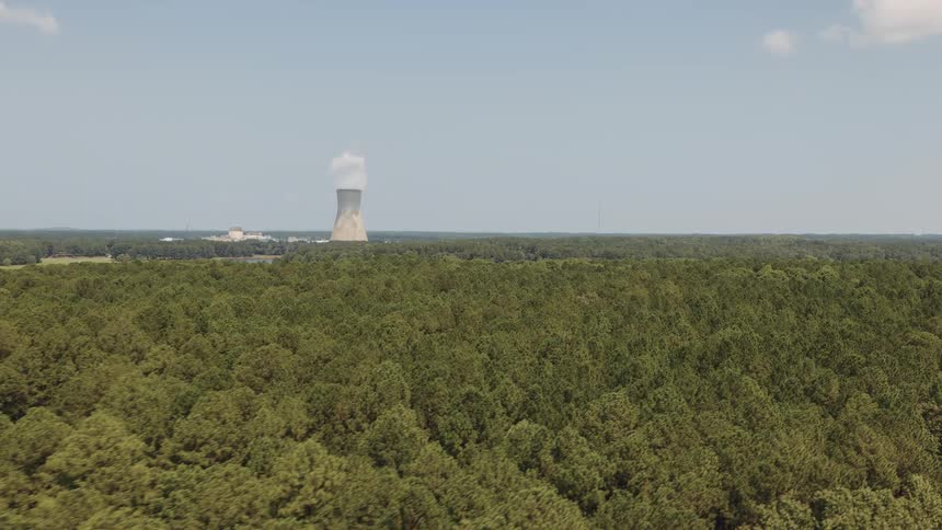 A slow aerial pullback reveals a nuclear power plant with its cooling tower steaming above a vast, lush forest