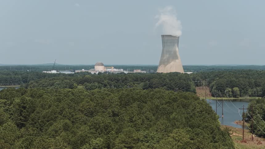 A slow aerial dolly shot pushes over a lush green forest towards a nuclear power plant's cooling tower emitting steam