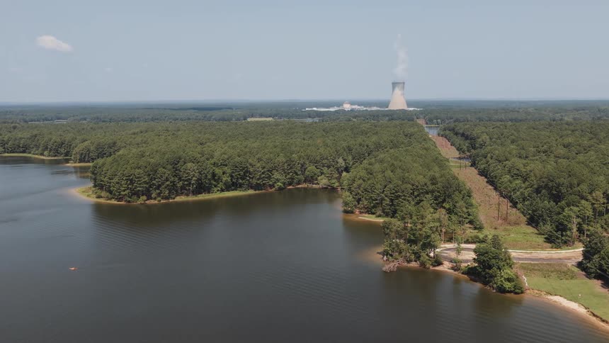 A wide aerial shot reveals a steaming nuclear power plant cooling tower situated by a serene lake and dense forest