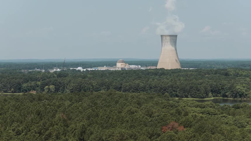 An aerial view reveals a nuclear power plant's cooling tower emitting steam, nestled within a vast forest next to a serene lake