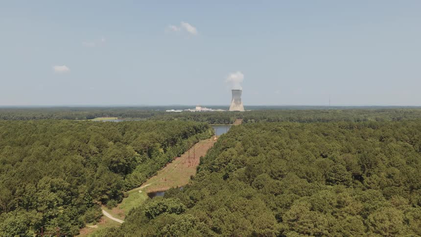A slow aerial push-in shot of a nuclear power plant and its cooling tower nestled in a dense forest next to a large lake on a sunny day