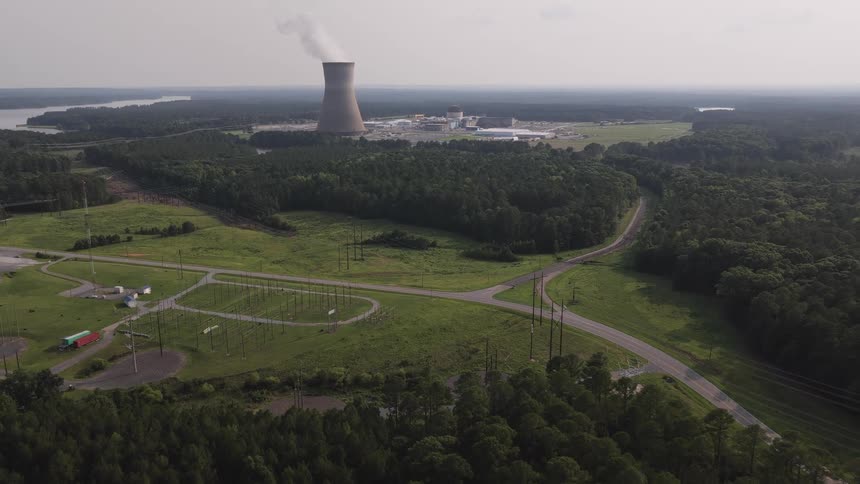 Wide aerial shot slowly retreats from a nuclear power plant with a smoking cooling tower, surrounded by a dense green forest and electrical grid