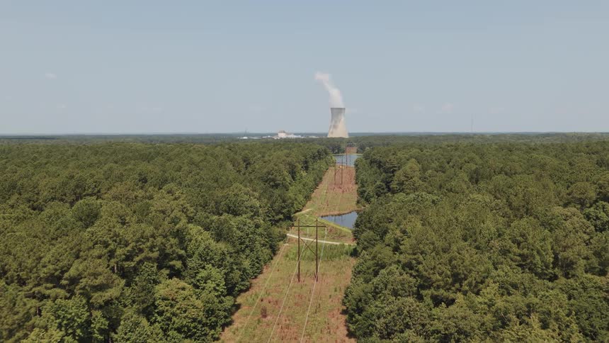 Aerial view flying over a lush green forest along power lines towards a nuclear power plant's cooling tower emitting steam