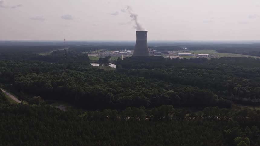 A slow-pulling back aerial view reveals a nuclear power plant's cooling tower emitting steam amidst a vast green forest and a nearby river