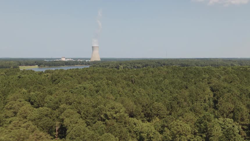 Aerial view flying over a dense green forest towards a nuclear power plant cooling tower on a sunny day