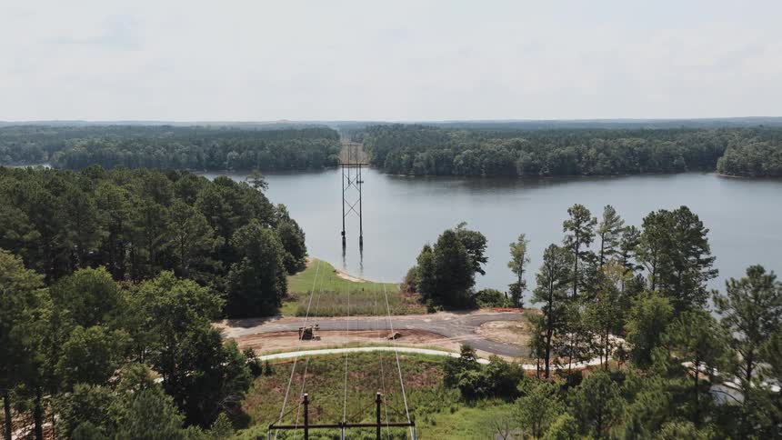 An aerial view shows power lines cutting a path through a dense forest and across a wide, calm lake