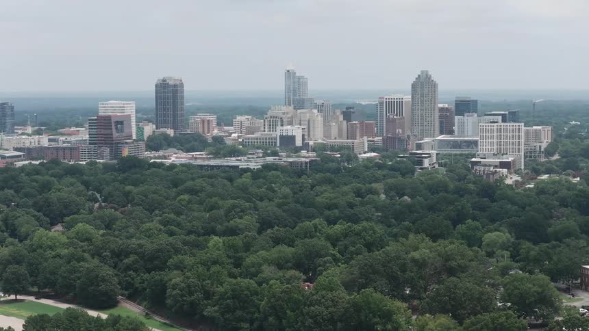 A slow-moving aerial shot captures the city skyline of Raleigh, North Carolina behind a lush green park on an overcast day
