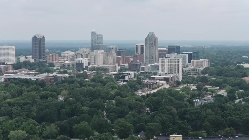A slow aerial pullback reveals the downtown Raleigh, North Carolina skyline surrounded by a lush green forest on a cloudy day