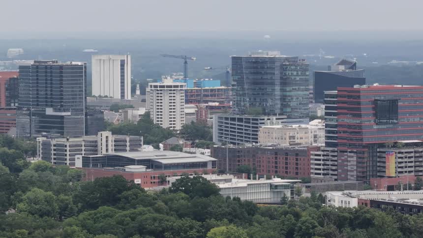 Slow aerial pan across a modern city skyline with diverse architecture surrounded by lush green trees on an overcast day