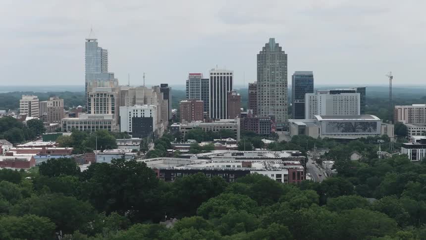 Aerial footage slowly pans right to reveal the downtown Raleigh, North Carolina skyline rising above a lush green forest on an overcast day