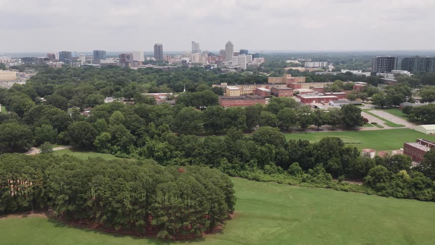 A stationary aerial view shows a lush green park with the modern skyline of Raleigh, North Carolina in the background under a cloudy sky