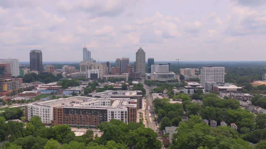 Aerial view slowly pushes in on the Raleigh, North Carolina skyline over lush green trees on a partly cloudy day