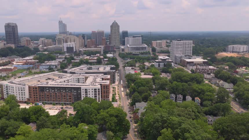 A slow aerial push-in shot revealing the Raleigh, North Carolina skyline over lush green trees and residential neighborhoods on a partly cloudy day