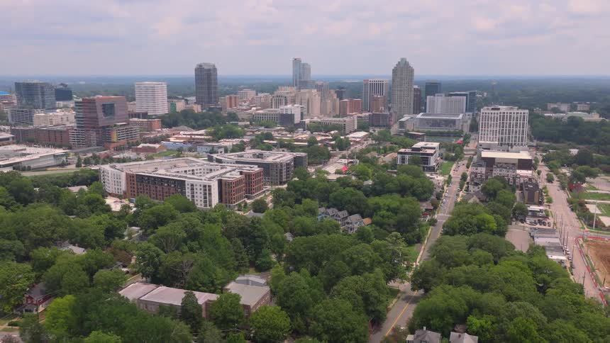 A slow aerial pullback reveals the modern city skyline of Raleigh, North Carolina, rising above a lush, green urban forest on a hazy day