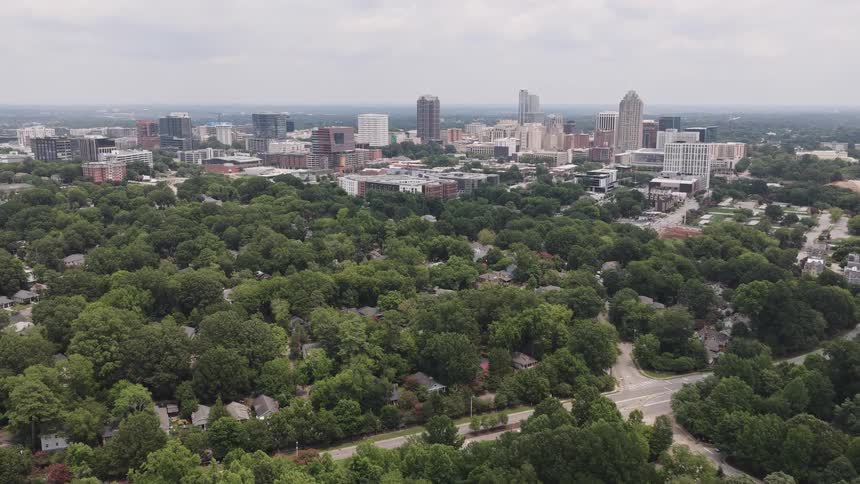 Aerial view slowly pushes over a lush, tree-filled park toward the Raleigh city skyline