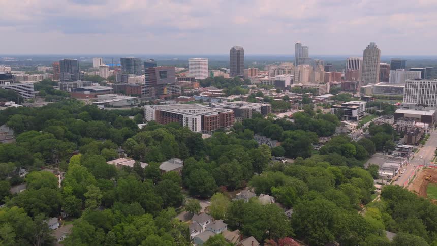 Slow aerial pullback reveals the lush green canopy of a residential neighborhood with the downtown Raleigh, North Carolina skyline in the background