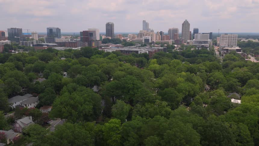 A slow aerial pullback reveals the lush green trees of a quiet neighborhood with the modern downtown Raleigh, North Carolina skyline in the background