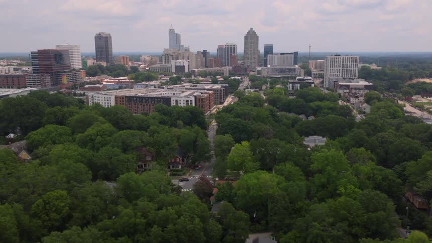 A slow aerial push-in shot over a lush, green neighborhood with the downtown Raleigh, North Carolina skyline in the background