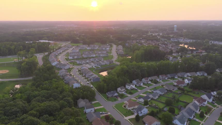 An aerial view captures a quiet suburban neighborhood with cookie-cutter houses nestled among lush green trees during a beautiful golden sunset
