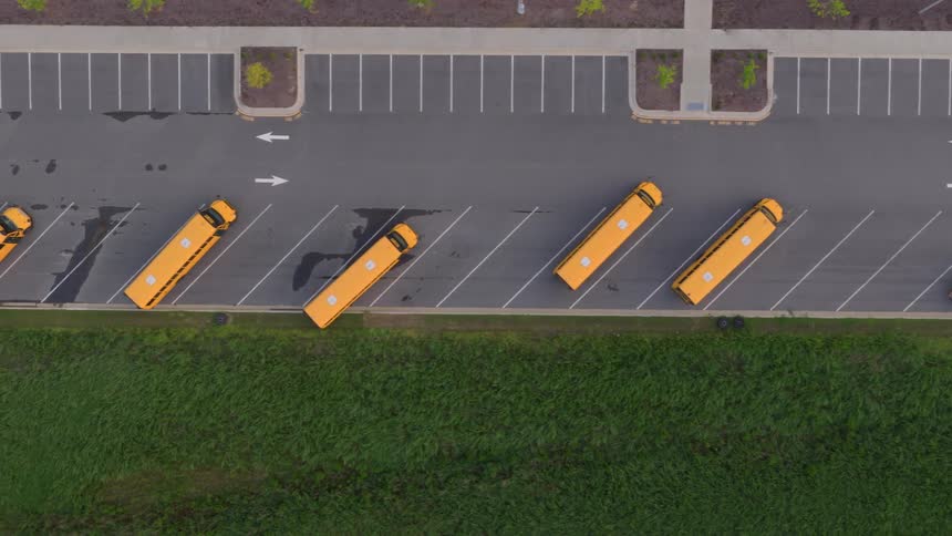 A slow ascending top-down aerial view of yellow school buses neatly parked in a large, empty parking lot next to a green field