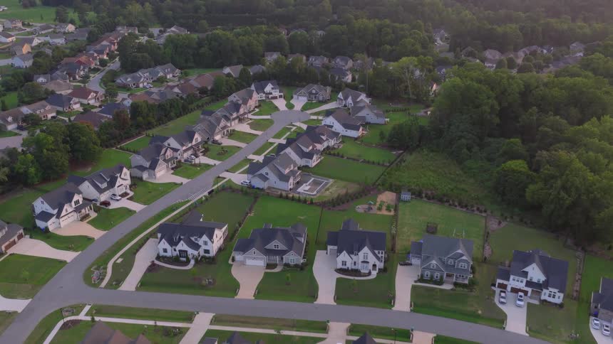 An aerial view slowly advances over a modern suburban neighborhood with large homes, green yards, and a cul-de-sac during golden hour