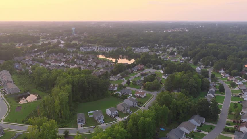 A slow aerial view reveals a quiet suburban American neighborhood with a tranquil lake and lush green trees during a hazy sunset