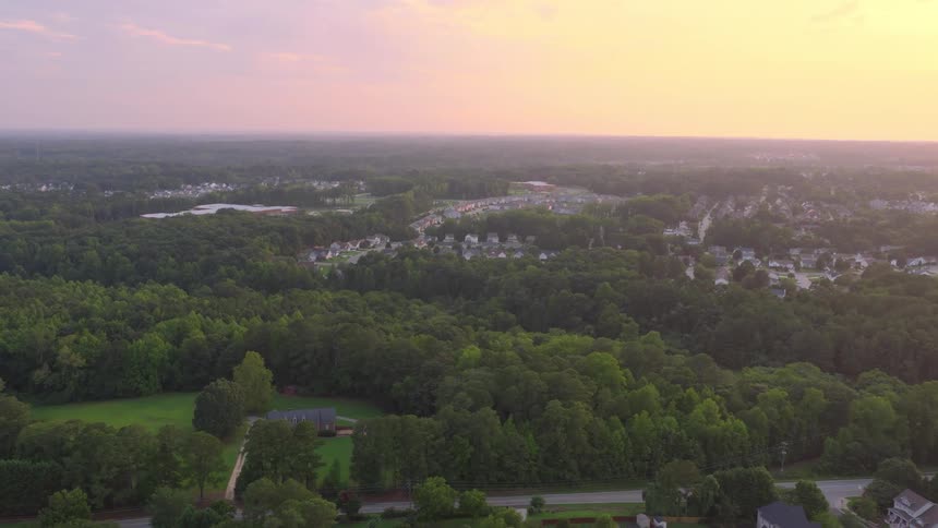Aerial footage captures a peaceful suburban neighborhood surrounded by lush green forests under a beautiful sunset sky