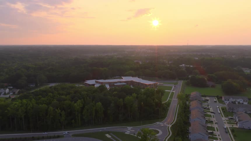 An expansive aerial view slowly approaches a school nestled in a forest, surrounded by a suburban community at sunrise