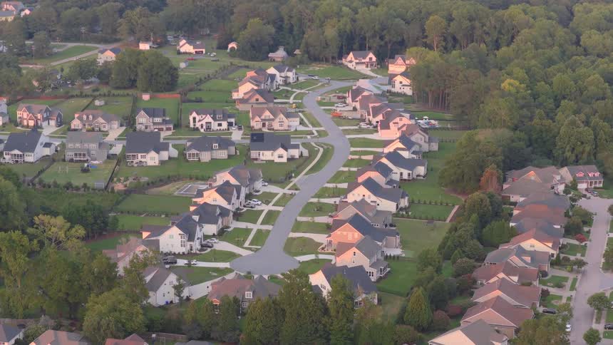 An aerial time-lapse pulls back to reveal a tranquil suburban neighborhood as the sun sets, casting long shadows across modern homes and green lawns