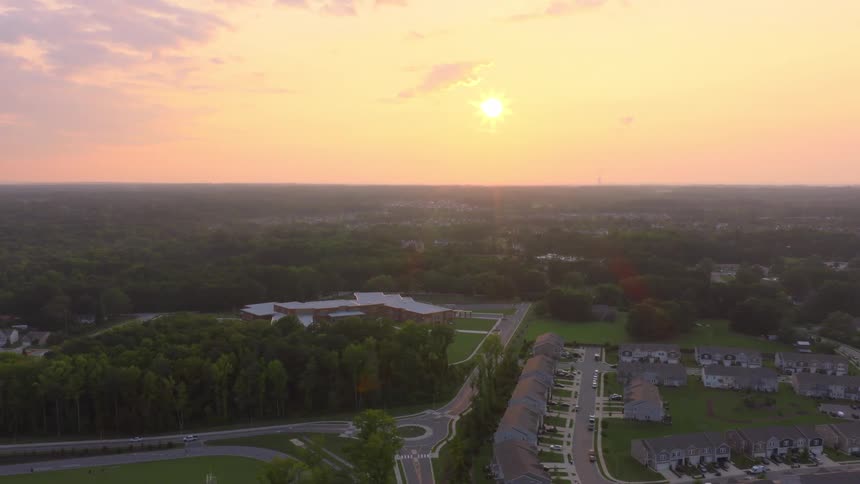 A hazy sunrise casts a warm glow over a suburban American neighborhood with a school and streets surrounded by dense forest