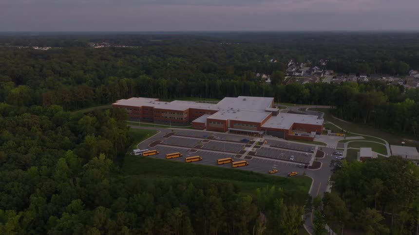 A tranquil aerial view pulls back from a large, modern school building with parked school buses, surrounded by a vast green forest at dusk