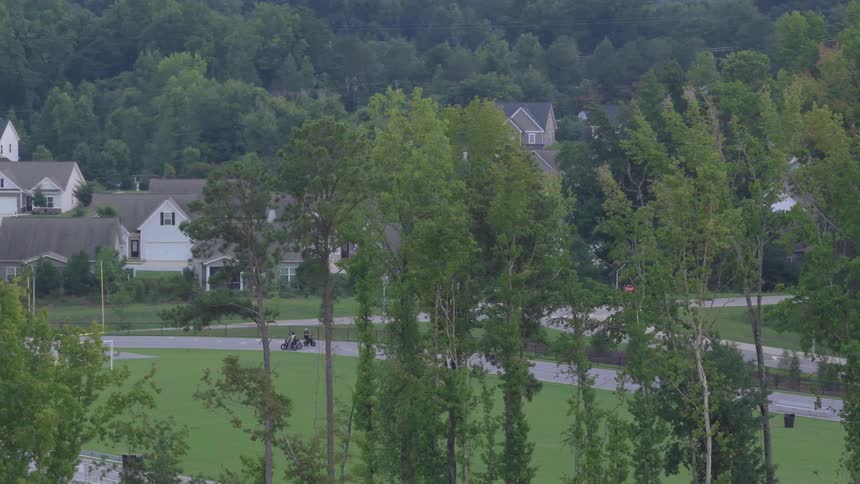A slow zoom-out shot overlooking a lush, wooded suburban neighborhood with a sports field and passing traffic