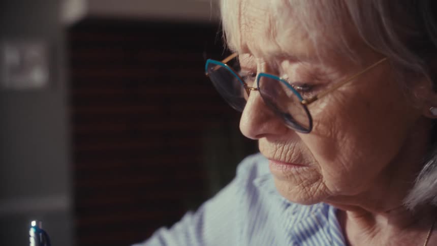 A focused senior woman with glasses writes a reminder on a sticky note while organizing paperwork at her desk