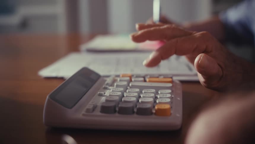 Close-up of a senior's hand using a calculator to manage finances, with another person writing in the background