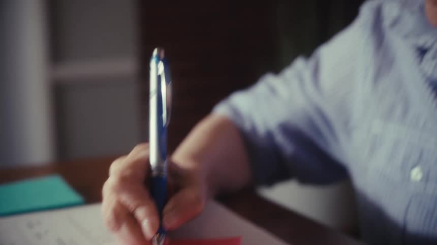 A focused elderly woman with glasses writes a note on a sticky pad while organizing financial documents and bills at her desk