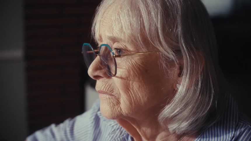 An intimate close-up profile of a thoughtful elderly woman with grey hair and glasses, sitting quietly indoors