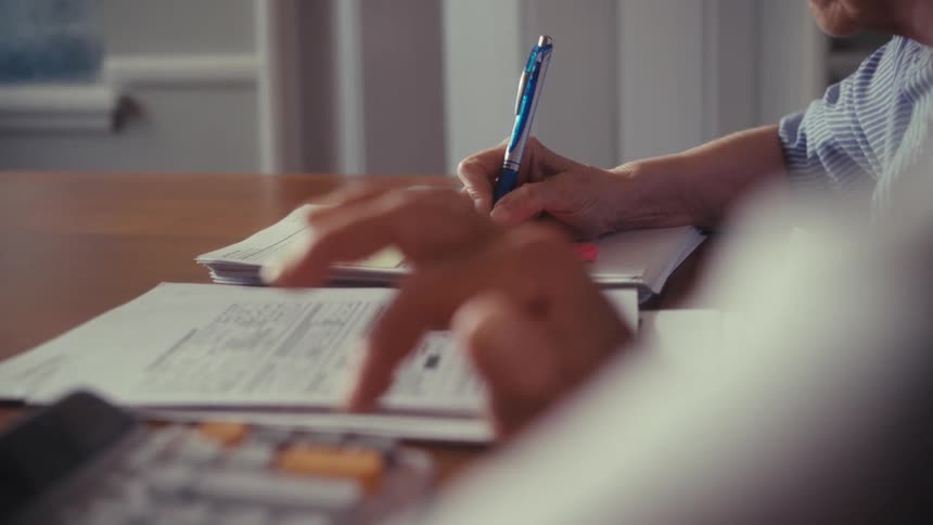 An elderly couple works together on their finances at a wooden table, one using a calculator and the other writing notes