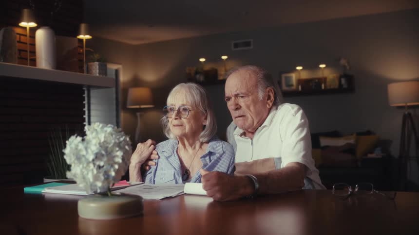 A worried senior couple sits together at their dining table at night, looking at something off-screen with concern