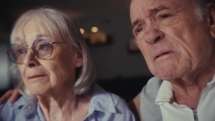 A close-up of an elderly couple with anxious and worried expressions watching something intently together at home