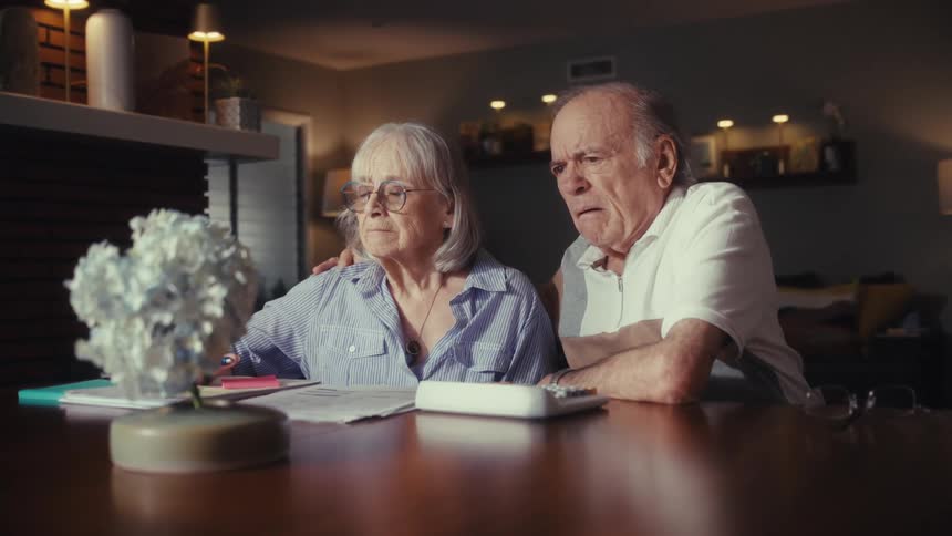 An elderly couple with worried expressions sit at their dining table at night looking at their finances