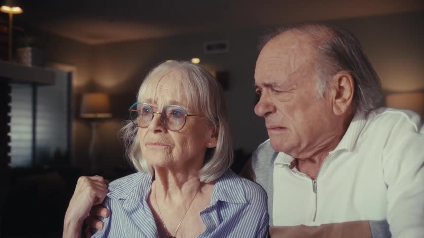 An elderly couple with concerned expressions watch something troubling on a screen in their home at night