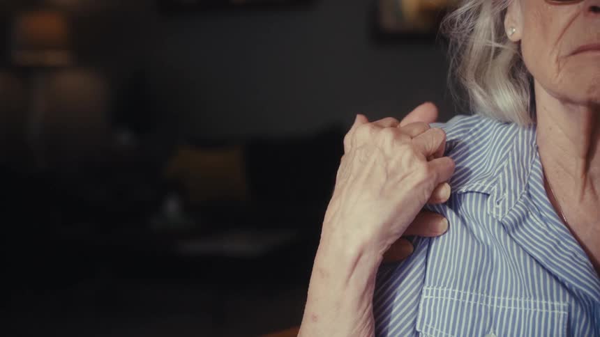 A supportive hand rests on a worried elderly woman's shoulder, and she holds it in a tender gesture of comfort and connection