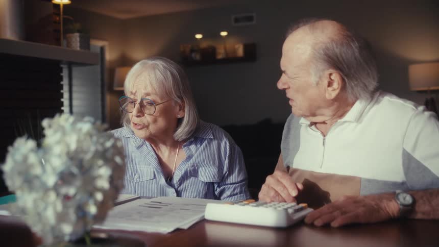 A worried elderly couple sits at a table in their home at night, discussing their finances and calculating bills