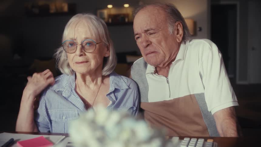 A worried senior couple sits at their dining room table going over bills and finances, comforting each other