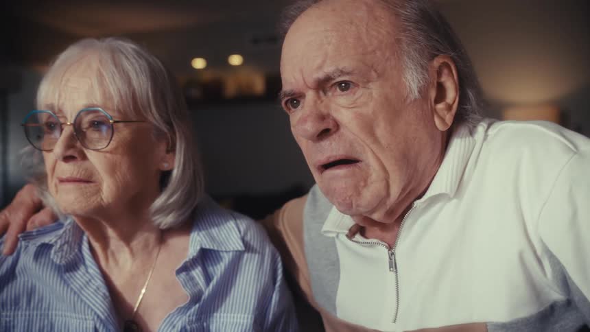 A worried elderly couple sits at their dining table calculating bills, their expressions turning to shock and despair over their financial situation