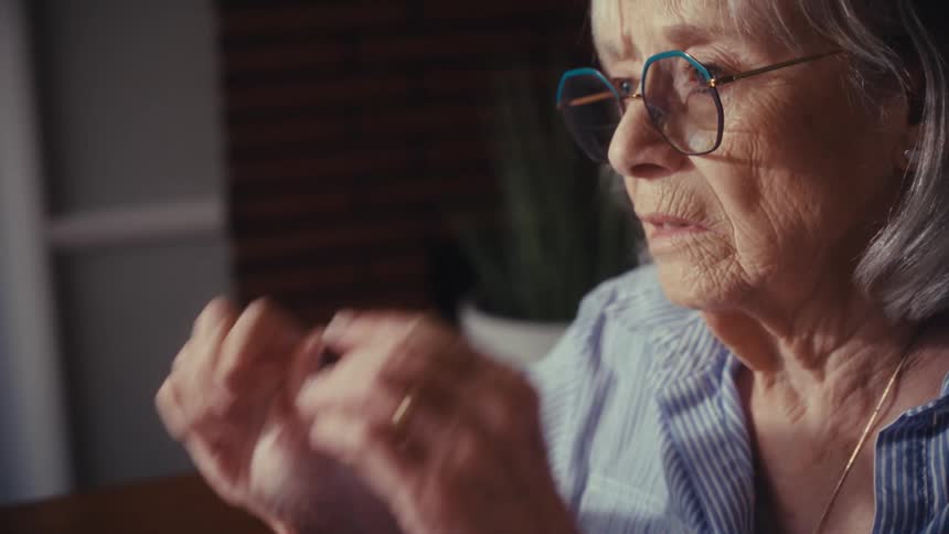 A stressed elderly woman writes a 'Past Due!!' note while sorting through bills, then takes off her glasses and looks away with a worried expression